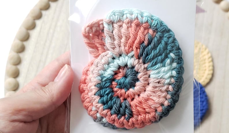 May include: A crocheted seashell in shades of pink, teal, and white. The seashell is a spiral shape and is made with a textured stitch.