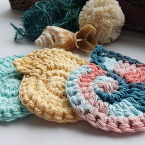 May include: Four crocheted cotton scrubbies in different colors and patterns. The scrubbies are in the shape of a circle and have a textured surface. The colors are light blue, yellow, pink, and blue.
