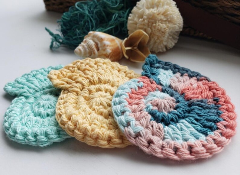 May include: Four crocheted cotton scrubbies in different colors and patterns. The scrubbies are in the shape of a circle and have a textured surface. The colors are light blue, yellow, pink, and blue.