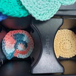 May include: Three crocheted cup coasters in a car cup holder. The coasters are in a spiral pattern and are in shades of blue, pink, and beige.