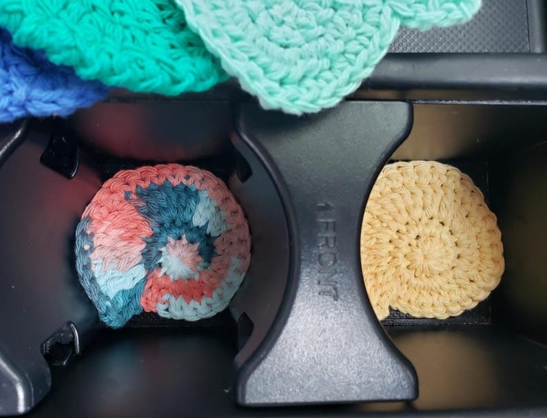 May include: Three crocheted cup coasters in a car cup holder. The coasters are in a spiral pattern and are in shades of blue, pink, and beige.