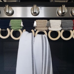 May include: Eight crocheted pot holders in various colors, including yellow, green, black, white, gray, and red, are hanging on a stovetop rack with wooden rings. Two white kitchen towels with gray stripes are hanging on the rings.