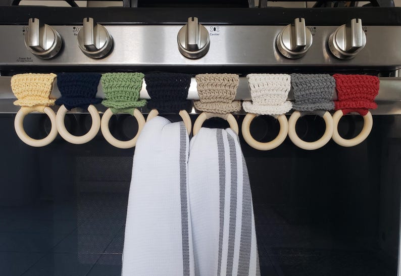 May include: Eight crocheted pot holders in various colors, including yellow, green, black, white, gray, and red, are hanging on a stovetop rack with wooden rings. Two white kitchen towels with gray stripes are hanging on the rings.