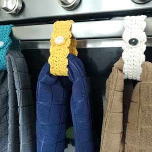 Pode incluir: Tr&ecirc;s porta-toalhas de croch&ecirc; em branco, amarelo e azul-turquesa, pendurados na ma&ccedil;aneta de uma porta de forno. Cada porta-toalhas tem um bot&atilde;o no centro. Os porta-toalhas est&atilde;o segurando toalhas em marrom, azul e cinza.
