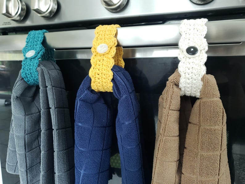 Pode incluir: Tr&ecirc;s porta-toalhas de croch&ecirc; em branco, amarelo e azul-turquesa, pendurados na ma&ccedil;aneta de uma porta de forno. Cada porta-toalhas tem um bot&atilde;o no centro. Os porta-toalhas est&atilde;o segurando toalhas em marrom, azul e cinza.