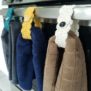 Pode incluir: Quatro toalhas de cozinha penduradas em um rack de forno de prata. As toalhas s&atilde;o azul marinho, marrom, branco e amarelo. Cada toalha tem um la&ccedil;o de croch&ecirc; com um bot&atilde;o.