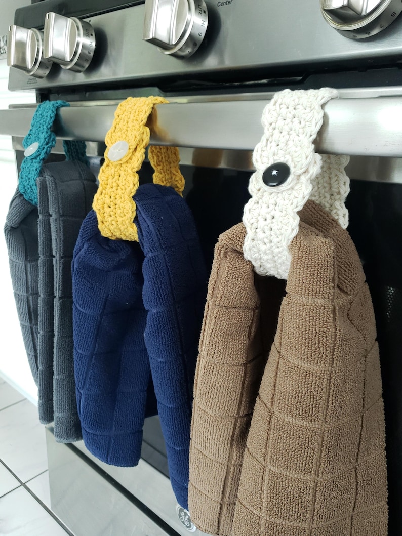 Pode incluir: Quatro toalhas de cozinha penduradas em um rack de forno de prata. As toalhas s&atilde;o azul marinho, marrom, branco e amarelo. Cada toalha tem um la&ccedil;o de croch&ecirc; com um bot&atilde;o.