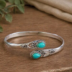 May include: A silver cuff bracelet featuring an open design with two teardrop-shaped turquoise stones at the ends. The metalwork surrounding each stone displays intricate filigree patterns and small beaded details along the band edges.