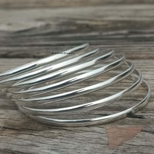 May include: A stack of six shiny silver bangles, each with a smooth, rounded design. The bangles are arranged on a rustic wooden surface, creating a contrast between the polished metal and the natural wood grain. The jewelry is reflective.