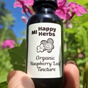 May include: A dark amber glass dropper bottle with a black cap and a white label. The label features the text MI Happy Herbs, a small flower graphic, and the words Organic Raspberry Leaf Tincture in black ink against a plain white background.