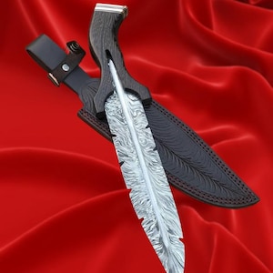 Puede incluir: Cuchillo artesanal con una hoja en forma de pluma y un mango de madera oscura. La hoja tiene un diseño único. El cuchillo está en una funda de cuero negro con trabilla, sobre un fondo rojo.