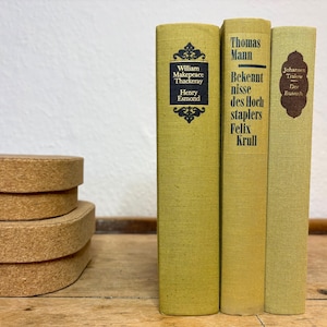 Könnte beinhalten: Drei alte Bücher mit gelben Einbänden werden auf einer Holzoberfläche präsentiert. Die Bücher haben Titel und Autorennamen auf ihren Buchrücken aufgedruckt. Ein Stapel Untersetzer aus Kork befindet sich im Vordergrund. Die Bücher sind in gutem Zustand.