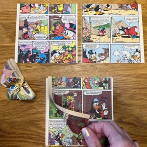 Peut inclure: Trois pages de bande dessinée mettant en scène Donald Duck dans des scènes colorées variées. Les pages sont disposées sur une surface en bois. Une page est partiellement pliée.