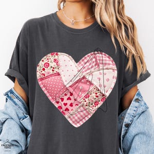 Puede incluir: Camiseta gris oscuro con un diseño de corazón de patchwork. El corazón está compuesto por varias telas rosas, incluyendo lunares, estampados florales y cuadros vichy. Un dibujo lineal negro de una figura está cosido encima del corazón.