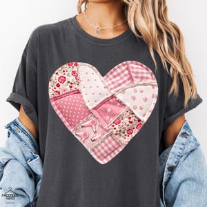 May include: Dark gray t-shirt featuring a patchwork heart design. The heart is composed of various pink fabrics, including floral, polka dot, and gingham patterns. Pink hair dryers are also featured in the design. The shirt has rolled-up sleeves.