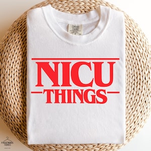 May include: White t-shirt with the red text "NICU THINGS" in a bold, stylized font. The shirt is made of a soft material and has a crew neck. The shirt is displayed on a woven, circular surface.