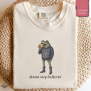 May include: Cream-coloured t-shirt with a cartoon frog wearing a blue sweater with a menorah design, purple boots, and eating a doughnut. The text "donut stop believin'" is printed below the frog. The shirt has a Comfort Colors tag.