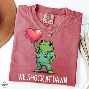 Könnte beinhalten: Ein altrosa T-Shirt mit einer Grafik eines grünen Frosches in medizinischer Kleidung mit Stethoskop, der ein rosa Herz hält. Unter dem Frosch steht der Text WE SHOCK AT DAWN. Das T-Shirt hat ein Comfort Colors Etikett am inneren Halsausschnitt.