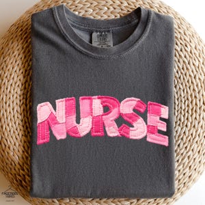 Puede incluir: Una camiseta gris oscuro con la palabra "NURSE" en letras rosas de patchwork en la parte delantera. Las letras son de diferentes tonos de rosa y están cosidas a la camiseta. La camiseta está doblada sobre una superficie tejida.