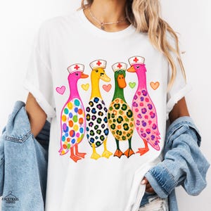 Könnte beinhalten: Ein weißes T-Shirt mit einer Grafik von vier Enten, die Krankenschwesterhauben mit roten Kreuzen tragen. Die Enten haben bunte Muster wie Punkte, Leopardenmuster und Flecken. Zwischen den Enten befinden sich kleine bunte Herzen.