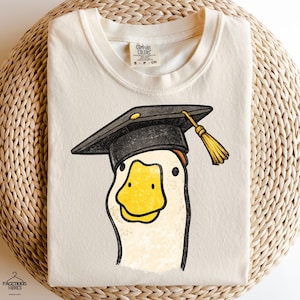 Könnte beinhalten: Cremefarbenes T-Shirt mit einer Cartoon-Gans, die einen schwarzen Abschluss-Hut mit einer goldenen Quaste trägt. Die Gans hat einen gelben Schnabel und lächelt. Das T-Shirt ist auf einer gewebten Oberfläche gefaltet.