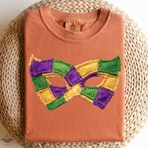 以下が含まれることがあります： 鮮やかなマルディグラマスクのデザインが施された、オレンジ色のTシャツ。マスクは緑、金、紫のパッチワークの正方形でできており、縁がほつれています。シャツにはComfort Colorsのタグが付いています。
