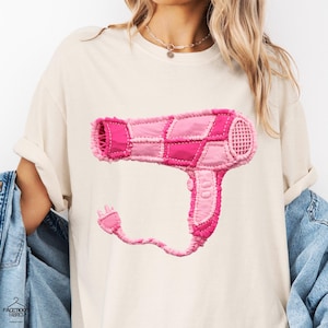 Puede incluir: Camiseta color crema con un diseño de patchwork rosa de un secador de pelo. El gráfico del secador de pelo incluye varios tonos de rosa y un enchufe. La camiseta tiene mangas cortas y un corte holgado.