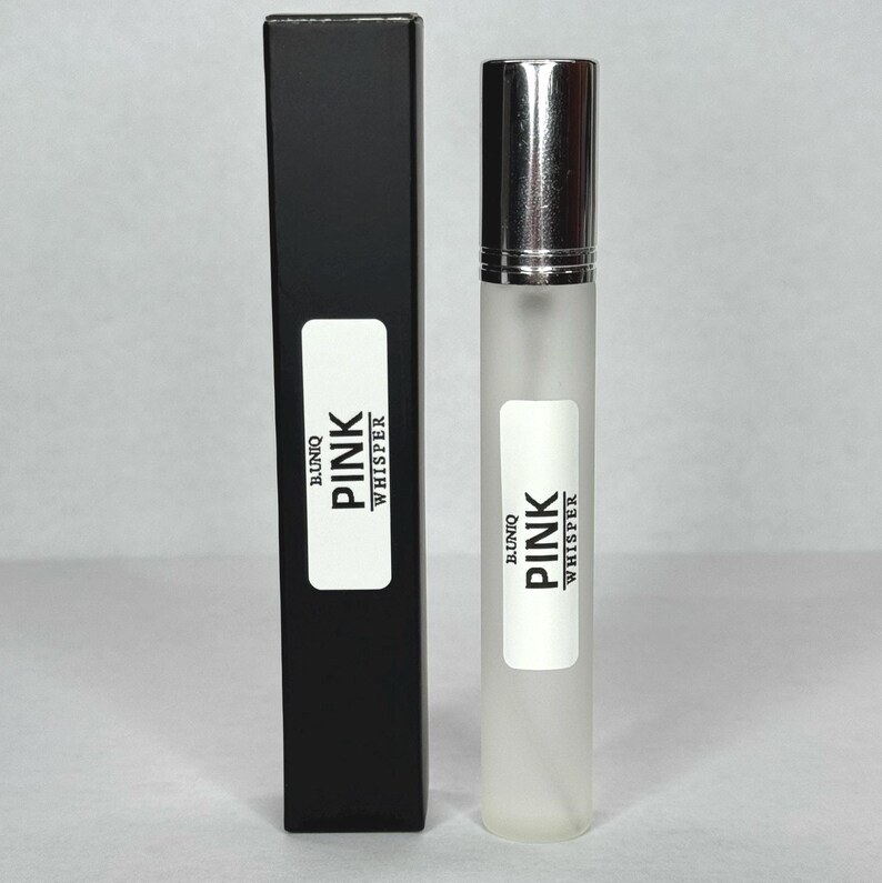 May include: A perfume bottle and its black box are displayed. The bottle is clear with a silver cap and a white label that reads "BUNIQ PINK WHISPER." The box has a matching label. The background is a neutral color.