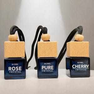 May include: Three dark blue, square-shaped car air fresheners with wooden tops and black cords. Each bottle has a different scent name in white text: Rose Crystal, Pure Crystal, and Cherry Crystal. The brand name B.UNIQ is also visible.