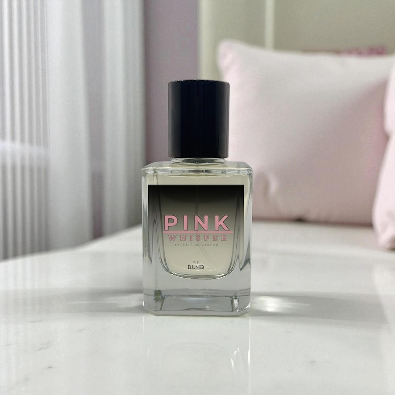 May include: A clear glass perfume bottle with a black cap. The label reads "PINK WHISPER" in pink and white text. The bottle is rectangular and sits on a white surface.