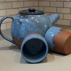 May include: A handmade ceramic tea set in shades of blue and brown. The set includes a teapot with a lid and two matching cups. The teapot and cups have a textured, glazed finish. The set is designed for serving tea.