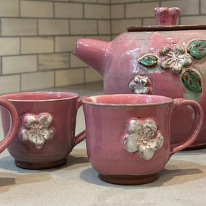 May include: A pink ceramic tea set featuring a teapot, mugs, and a small lidded dish. Each piece is adorned with white and green floral accents. The set is designed for serving tea.