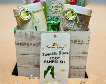 May include: A white wooden box filled with a foot pamper kit. The kit includes a pair of green socks, a small bag of tea, a nail file, and a small bottle of bath salts. The box has a tag that says "Lemon Drop Gift Shop Twinkle Toes Foot Pamper Kit".