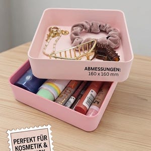 Może przedstawiać: Dwa, różowe, piętrowe pojemniki do przechowywania, każdy o wymiarach 16 x 16 cm, wypełnione kosmetykami i akcesoriami. Górny pojemnik zawiera gumkę do włosów, spinkę i biżuterię. Dolny pojemnik zawiera kosmetyki.