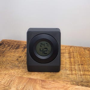 Puede incluir: Un termómetro e higrómetro digital negro muestra 24,7 grados Celsius y 42% de humedad. El dispositivo, de forma cuadrada, tiene una pantalla circular y está sobre una superficie de madera.