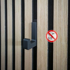 Könnte beinhalten: Ein mattschwarzer J-förmiger Haken, der an einer vertikalen Holzlattenwand befestigt ist. Rechts befindet sich ein kreisförmiges rot-weißes Symbol mit einer durchgestrichenen Bohrmaschine, das anzeigt, dass kein Bohren für die Montage nötig ist.