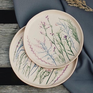 Pode incluir: Dois pratos de cerâmica creme com um design botânico. Os pratos apresentam ilustrações delicadas de flores silvestres em tons de roxo, verde e azul. Os pratos estão empilhados um sobre o outro.