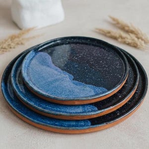 May include: A stack of three ceramic plates with a unique design. The plates feature a dark blue and speckled finish, with a lighter blue wave pattern. The rims are a natural terracotta color, creating a contrast. These plates are ideal for serving food.