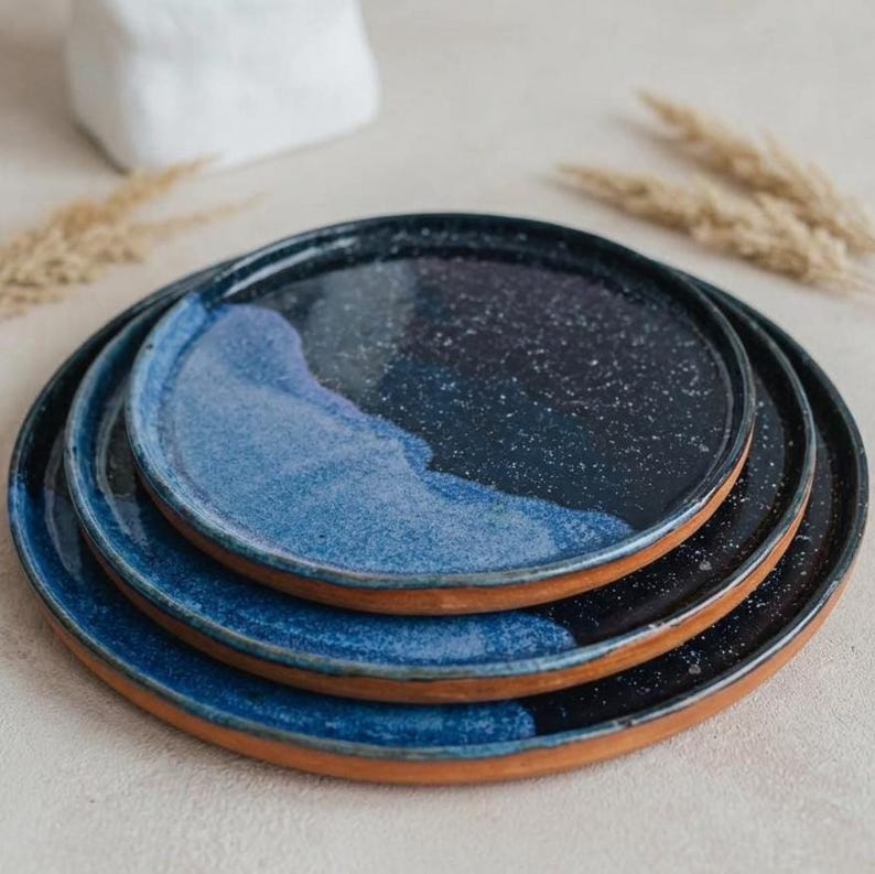 May include: A stack of three ceramic plates with a unique design. The plates feature a dark blue and speckled finish, with a lighter blue wave pattern. The rims are a natural terracotta color, creating a contrast. These plates are ideal for serving food.