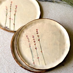 May include: Two cream-coloured ceramic plates with a brown rim and lavender sprig design. The lavender features pink flowers and green stems. The plates are on a textured, light-coloured surface.