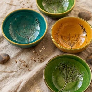 May include: Three ceramic bowls in teal, mustard yellow, and light green, each featuring a black botanical illustration of a seed head or flower. The bowls have smooth, rounded interiors and are arranged on a textured fabric surface.
