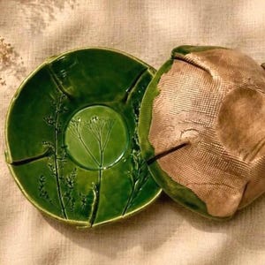 Könnte beinhalten: Eine runde Keramikschale mit glänzender grüner Glasur und einem geprägten botanischen Pflanzendesign. Daneben liegt ein passender Keramikdeckel mit strukturierter, goldener Metallic-Oberfläche und einem gitterartigen Muster.