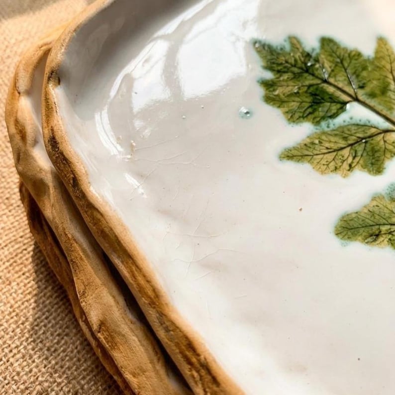 May include: A stack of handmade ceramic plates. The plates have a white glazed surface with a green leaf design. The edges are a natural brown colour, giving them a rustic appearance. The plates are perfect for serving food.