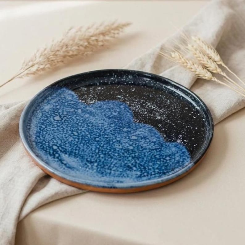 May include: A round ceramic plate with a speckled blue and black design. The plate has a wavy blue pattern on the left side and a black speckled pattern on the right. The rim is a natural terracotta color.