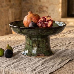 May include: A pedestal bowl with a glossy, mottled dark green glaze. The bowl contains one whole pomegranate, one halved pomegranate showing red seeds, and one dark purple fig. A small stamp is visible on the base of the ceramic pedestal bowl.