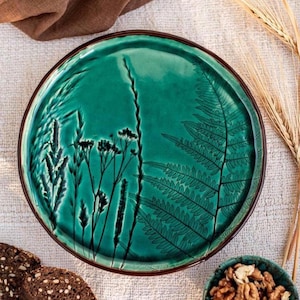 May include: A teal ceramic plate with a brown rim, featuring a botanical design of ferns and grasses. The plate has a glossy finish and is surrounded by wheat stalks, bread, and a bowl of walnuts.