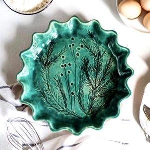 May include: A round ceramic pie dish with a ruffled edge and a glossy emerald green glaze. The interior features a dark, etched botanical design of pine branches and small berries centered on the base, creating a textured, detailed appearance.