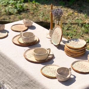 May include: A collection of handmade ceramic tableware, including cups, saucers, and plates, arranged on a white fringed tablecloth. The pottery has a rustic, textured appearance with a cream color and brown accents. A vase of lavender and a candle are also present.