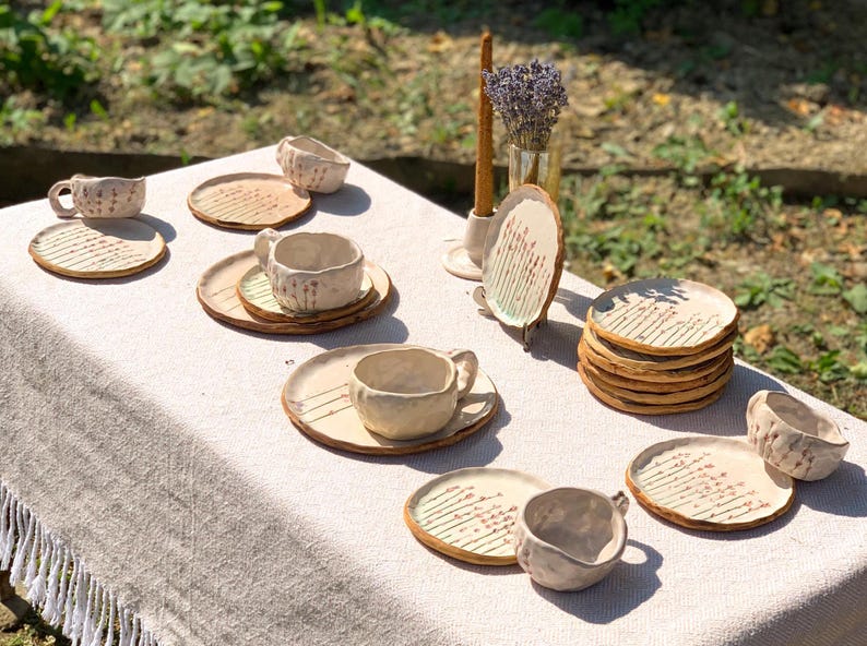 May include: A collection of handmade ceramic tableware, including cups, saucers, and plates, arranged on a white fringed tablecloth. The pottery has a rustic, textured appearance with a cream color and brown accents. A vase of lavender and a candle are also present.