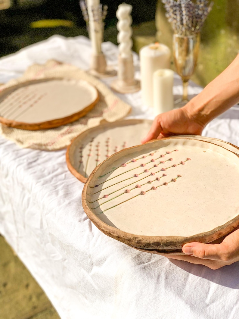 May include: A set of beige ceramic plates with a brown rim and a floral design. The plates are on a white tablecloth, with candles and lavender in the background. The plates are being held by a person.